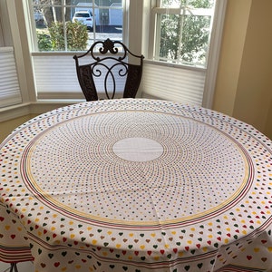 May include: A round tablecloth with a white background and a pattern of small hearts in red, yellow, and green. The tablecloth has a concentric circle design in the centre and is placed on a round table. A decorative chair is behind the table.