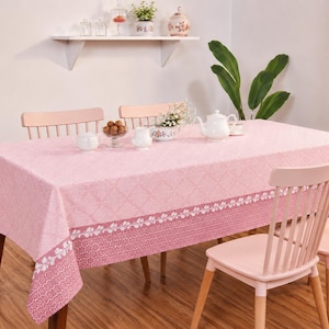 May include: A rectangular tablecloth in shades of pink featuring a repeating diamond pattern on the top surface and a darker pink border with a white floral vine motif along the edges, draped over a wooden table with matching pink chairs.