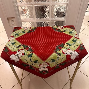 May include: A square red Christmas tablecloth with a green border featuring Santa Claus and holly designs. The tablecloth is placed on a gold-coloured metal table with four legs. The table is set against a white door with glass blocks.