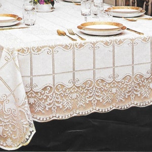 May include: A white lace tablecloth with a detailed floral and geometric pattern. The tablecloth is draped over a table set for a meal, with gold-rimmed plates, silverware, and glasses visible. The lace design features a scalloped edge.
