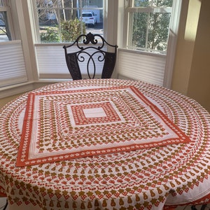 May include: A round table covered with a white tablecloth featuring a repeating pattern of red cherries, pears, and brown grapes. The tablecloth has a red border and is set on a dark metal chair. The table is in front of a window.
