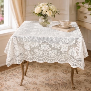 May include: A square white lace tablecloth featuring a detailed floral pattern with a scalloped edge. The tablecloth is draped over a table, topped with a clear glass vase containing white roses and a ceramic teacup resting on a book.