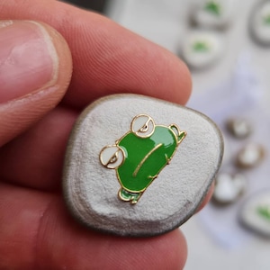 Op de afbeelding: Een kleine, ronde, grijze steen met een groene kikkerpin eraan vastgemaakt. De kikker kijkt naar rechts en heeft witte ogen.