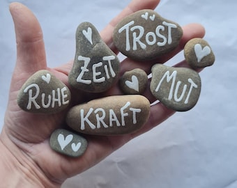 Könnte beinhalten: Eine Hand hält acht bemalte Steine mit den Worten "Ruhe", "Zeit", "Kraft", "Mut", "Trost" und Herzen.