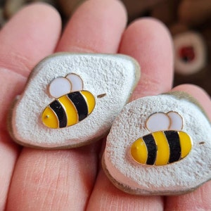 Könnte beinhalten: Zwei bemalte weiße Steine mit schwarzen und gelben Bienenmotiven. Die Bienen haben weiße Flügel.