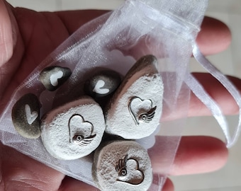Könnte beinhalten: Ein weißer Organzasack gefüllt mit kleinen grauen Steinen. Jeder Stein hat ein weißes Herz mit einem silbernen Flügeldesign darauf gemalt.