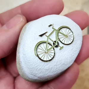 Puede incluir: Una piedra pintada de blanco con un diseño de bicicleta verde. La bicicleta tiene contornos negros y radios blancos.