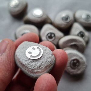 Könnte beinhalten: Eine Sammlung glatter, grauer Steine mit silbernen Smiley-Gesichtern, die in sie eingeätzt sind. Die Steine sind in verschiedenen Größen und Formen.