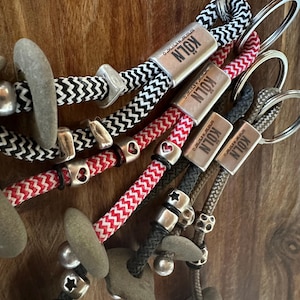 Op de afbeelding: Close-up van verschillende sleutelhangers met touwachtige koorden in zwart en wit, rood en wit, zwart en rood, zwart en groen en bruin en grijs. Elke sleutelhanger heeft een metalen label met "KÖLN" en een stenen hanger. De sleutelhangers liggen op een houten oppervlak.