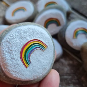 Op de afbeelding: Een verzameling beschilderde stenen met regenboogontwerpen. Elke steen is wit geschilderd met een kleurrijk regenboogontwerp.