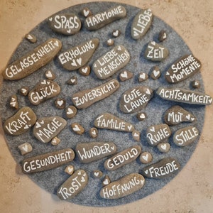 Peut inclure: Des galets bruns clairs avec des mots blancs et des cœurs blancs. Les mots incluent "Famille", "Espoir" et "Patience". Les pierres sont disposées sur un cercle de feutre gris.