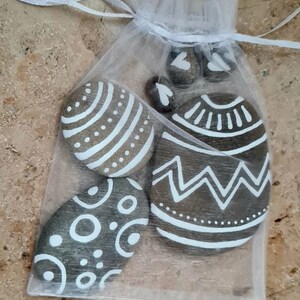 Könnte beinhalten: Ein Set aus fünf bemalten Steinen in einem weißen Organzasäckchen. Die Steine sind mit weißen geometrischen Mustern auf braunem Hintergrund bemalt.