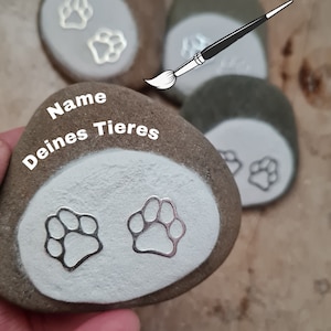 Könnte beinhalten: Drei graue Steine mit weiß bemalten Flächen. Die Steine haben silberne Pfotenabdrücke und den Text "Name deines Tieres" auf einem der Steine.