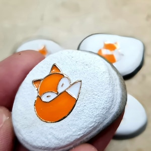 Könnte beinhalten: Eine Sammlung kleiner, weißer, strukturierter Steine, die jeweils mit einem leuchtend orangefarbenen und weißen Fuchsmotiv mit goldenen Konturen verziert sind. Die Steine sind glatt und rund, ideal für dekorative Zwecke.