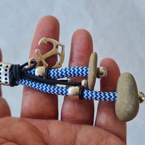 Op de afbeelding: Een blauwe en witte touw sleutelhanger met een zilveren ankerbedel, twee kleine grijze stenen en zilveren kralen.