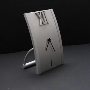 May include: A silver-coloured clock with Roman numeral markings on the face. The clock has a curved design and a stand that extends out from the bottom.