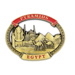 Może przedstawiać: Złoty metalowy magnes pamiątkowy przedstawiający szczegółowe przedstawienie piramid w Gizie w Egipcie. Magnes ma czerwoną wstęgę z napisem "PYRAMIDS EGYPT" i ozdobny brzeg w kształcie łańcucha.