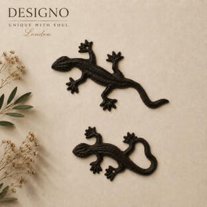 May include: Two black cast iron lizard-shaped wall hooks with textured surfaces. The top lizard is positioned horizontally with a straight tail, while the bottom lizard has a curved tail. Both feature visible screw holes for mounting purposes.