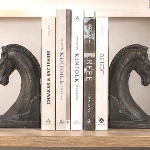 Può includere: Due fermalibri in ghisa grigio scuro a forma di teste di cavallo, rivolti verso l'interno di una fila di cinque libri. I libri hanno dorsi bianchi e grigio scuro con testo che include Kinfolk, Creee e altri titoli, tenuti in posizione.