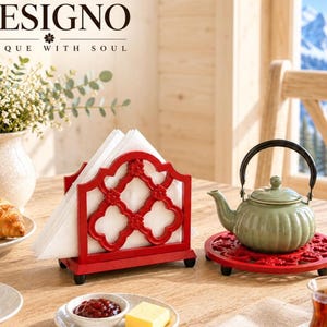 May include: A red metal napkin holder with a quatrefoil cutout pattern and small feet, holding white napkins. Beside it sits a light green ceramic teapot with a black handle on a matching red circular metal trivet with a floral cutout design.