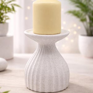 May include: A white ceramic candle holder with a textured, ribbed design. A pale yellow pillar candle sits atop the holder. The candle holder is on a wooden surface, with plants in the background.
