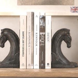 Peut inclure: Une paire de serre-livres en métal moulé gris foncé en forme de têtes de cheval, placés de chaque côté de cinq livres. Les livres incluent des titres comme Kinfolk, Creee et Abide, avec des dos présentant du texte noir sur fond blanc et gris clair.