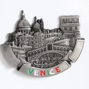 May include: A silver-coloured metal souvenir magnet depicting a detailed 3D model of the city of Venice, Italy. The magnet features a bridge, buildings, and a gondola. The word "VENICE" is written in red, white, and green on the bottom of the magnet.