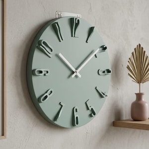 Pode incluir: Um relógio de parede redondo verde sálvia com ponteiros brancos e números estilizados em relevo. O relógio está montado numa parede cinza claro. Uma impressão botânica emoldurada e uma prateleira decorativa são visíveis.