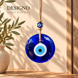 May include: A hanging wall ornament featuring a large, circular, deep blue glass evil eye charm with a central white and light blue eye design. It is suspended by a tan jute cord accented with a small square blue evil eye bead and gold-toned metallic spacers.
