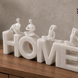 Può includere: Una scultura decorativa bianca che compone la parola HOME in lettere maiuscole. Ogni lettera presenta una piccola figura umana astratta bianca in una diversa posizione yoga. L'oggetto è realizzato in materiale liscio e opaco.