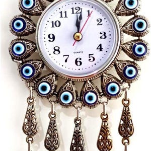 May include: A wall clock featuring a white face with black numerals and quartz movement, surrounded by a circular frame of blue and white evil eye beads in ornate metal settings, with dangling metal charms and a large blue glass evil eye pendant.