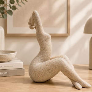 May include: A beige speckled abstract sculpture of a seated figure with arms raised above the head. The figure has a smooth, curved silhouette with legs extended forward and crossed at the ankles, crafted from a textured, stone-like material.