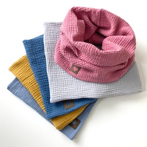 Könnte beinhalten: Ein Stapel aus fünf Stoffschals in verschiedenen Farben, darunter rosa, hellblau, dunkelblau und gelb. Jeder Schal hat ein kleines braunes Lederstück mit einem Stern- oder Herzmotiv.