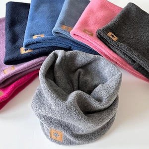 Könnte beinhalten: Ein Set aus sieben Fleece-Halswärmern in verschiedenen Farben, darunter Rosa, Lila, Blau und Grau. Jeder Halswärmer hat ein kleines Lederstück mit einem Sternmuster.