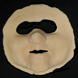 May include: A beige colored silicone face mask with a black eye patch and a detailed nose and mouth.