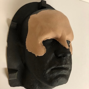 Puede incluir: Un molde negro de una cara humana con una máscara facial de arcilla de color tostado aplicada en la frente y la parte superior de las mejillas.