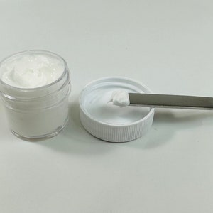 May include: A small white jar with a white lid and a white cream inside. A small amount of cream is on a white lid next to a silver spatula.
