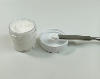 Op de afbeelding: Een klein wit potje met een wit deksel en een witte crème erin. Een kleine hoeveelheid crème bevindt zich op een wit deksel naast een zilveren spatel.