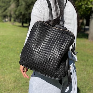 Könnte beinhalten: Ein schwarzer Rucksack mit einem geflochtenen Ledermuster auf der Außenseite. Die Tasche verfügt über einen Reißverschluss oben mit einem Lederzugband und zwei verstellbare Schultergurte. Das Design ist quadratisch mit abgerundeten Ecken.