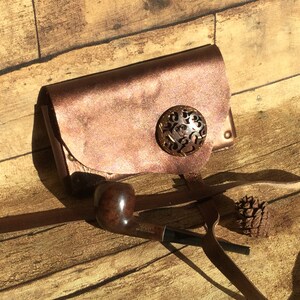 May include: A copper-colored leather pouch with a decorative metal button and a brown leather strap. A brown wooden pipe is lying on the strap.