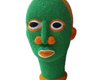 May include: A green beaded sculpture of a human head with orange and white details. The sculpture has a stylized face with large eyes, a nose, and a mouth.