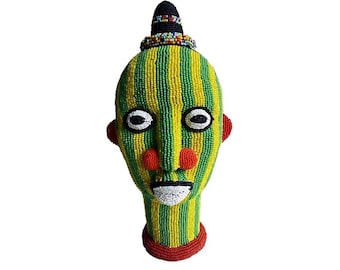 May include: A beaded human head sculpture with a green and yellow striped face, red lips, and red and white eyes. The sculpture has a black and white beaded crown on top of its head.