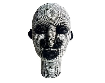 May include: A black and white beaded sculpture of a human head. The head is made of small, round beads that are arranged in a pattern to create the features of the face. The eyes, nose, and mouth are all clearly defined.