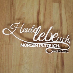 Könnte beinhalten: Ein weißes Holzschild mit dem Text "Heute lebe ich, Morgen putze ich. Vielleicht." Das Schild befindet sich auf einem Holzgrund.