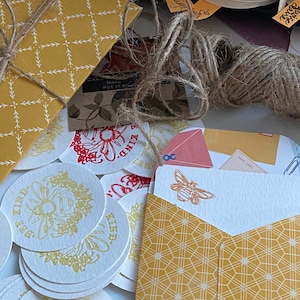 May include: A set of round paper coasters with a bee and flower design in yellow and white. The coasters are stacked on top of each other. There is a yellow envelope with a bee design and the text "Bee Kind" on it. The envelope is tied with twine.