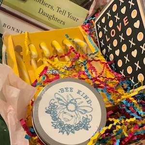 Könnte beinhalten: Ein Geschenkkorb gefüllt mit gelben und weißen gestreiften Zuckerstangen, eine kleine Dose mit einer Biene und den Worten "Bee Kind" und eine schwarz-weiße Karte mit dem Wort "LOVE" umgeben von Herzen und X.