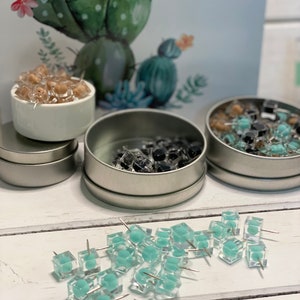 May include: Three metal tins filled with push pins. The tins are on a white wooden surface. The push pins are in various colors, including black, brown, and turquoise. A cactus illustration is in the background.