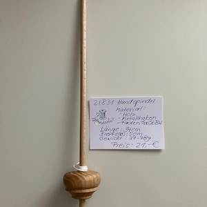 Op de afbeelding: Een houten handspindel met een afgeronde basis en een lange, slanke schacht. De spindel heeft een metalen ring aan de bovenkant en een punt. De tekst op het bijgevoegde papier zegt "21831 Handspindel". De spindel is 34 cm lang.