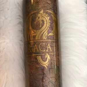 May include: A cylindrical tumbler with a textured, cracked-earth design in shades of brown, green, and gold. The tumbler features a gold dragon emblem and the word "DRACAR" in gold lettering, adding a fantasy theme.