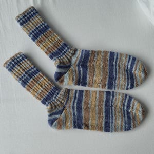 May include: A pair of knitted socks with a striped pattern in shades of blue, brown, and tan. The socks have a ribbed cuff and a striped design on the foot. The socks are displayed on a white surface.